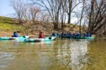 A group of people kayaking on a calm river, wearing life jackets. Bare trees and a grassy bank are visible in the background under a clear sky.