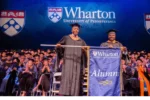 Photo of Wharton graduates in graduation regalia with two people in the front holding a Wharton alumni banner