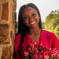 Adanna Mogbo smiling and holding pink flowers in a pink blazer posed against a red stone wall outside with trees in the background.