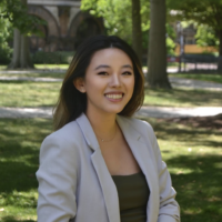 Alice Feng smiling in a dark green blouse and a light grey blazer outside in front of trees.