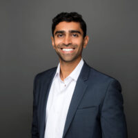 Aniket Patel smiling in a white button down and dark grey blazer in front of a dark grey background.