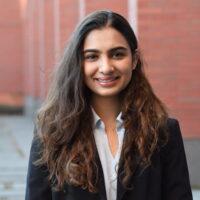 Bhavya Visal smiling in a white blouse and a black blazer in front of a red brick building.