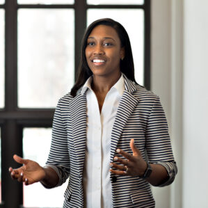 Brittney Jean-Louis smiling and talking in a black and white striped blazer over a white button down in front of a wall of windows.