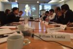 A group of people sits around a conference table during a meeting. Nameplates, notebooks, and cups are on the table. A projector displays a serious-looking individual on a screen.