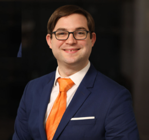 Evan Holownia smiling in a white button down, orange tie, and navy blue suit against a dark background.