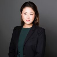 Jennie Zhang smiling in a dark green blouse and black blazer against a dark grey background.