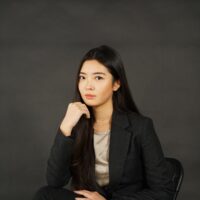 Louisa Qiu posed in a chair in a cream blouse and black suit in front of a dark grey background.