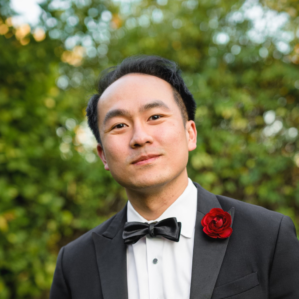 Michael Wong smiling in a black tuxedo, white button down, black bowtie, and red flower outside in front of foliage.