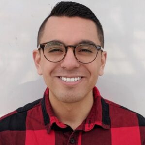 Richard Merino smiling in a red and black plaid button down against a cream wall.