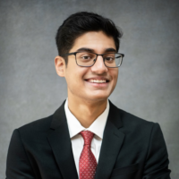 Roshan Pillai smiling in a white button down, red patterned tie, and a black blazer in front of a grey background.