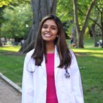 Media Sharma smiles in a bright pink dress and white coat with a stethoscope draped over her shoulders outside in front of grass and trees