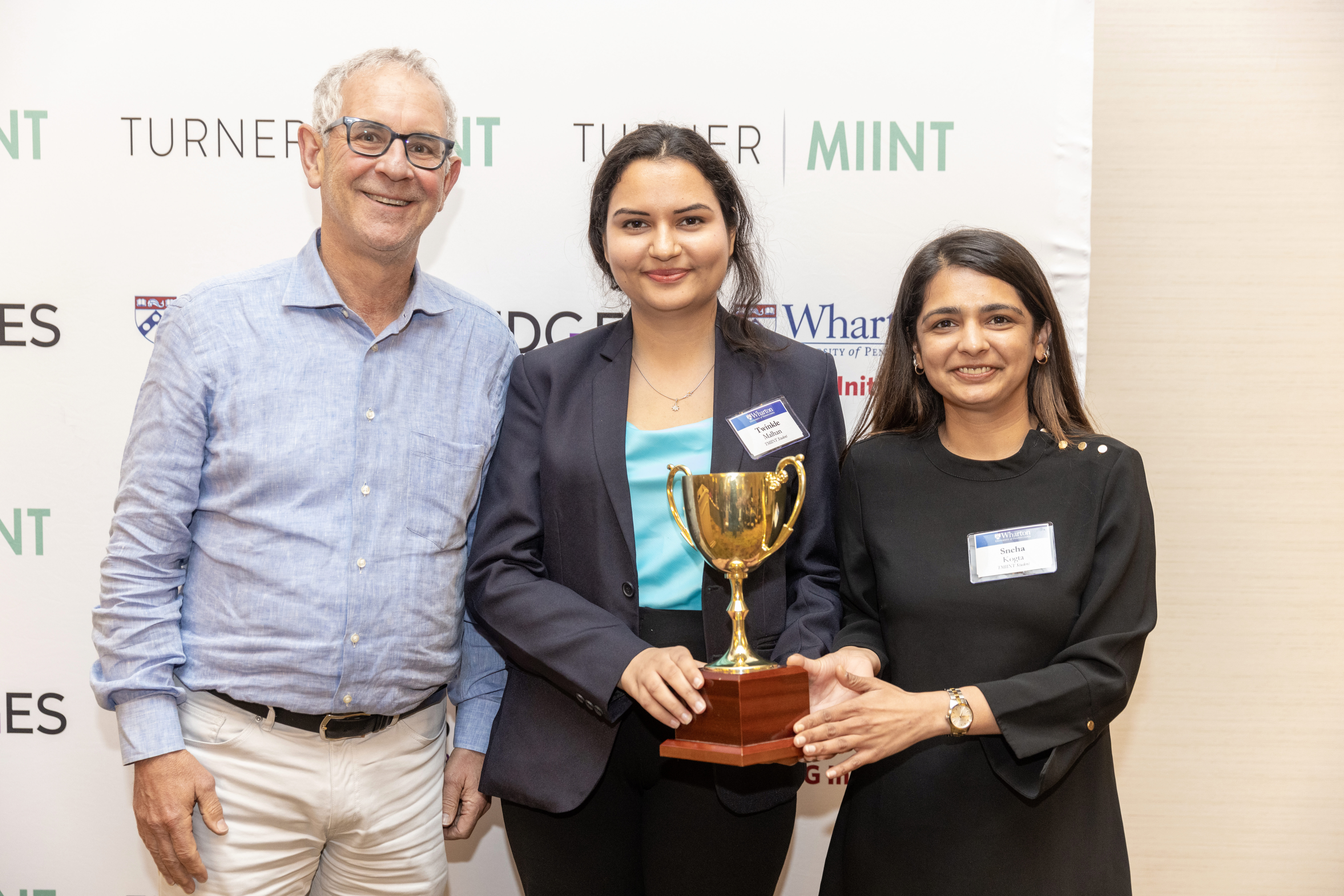 Ron Moelis poses with Twinkle Malhan and Sneha Kogta of the Saïd Business School Team. They're holding a gold tropy in front of a banner that says Turner MIINT, Wharton ESG Initiative, and Bridges Impact Foundation.