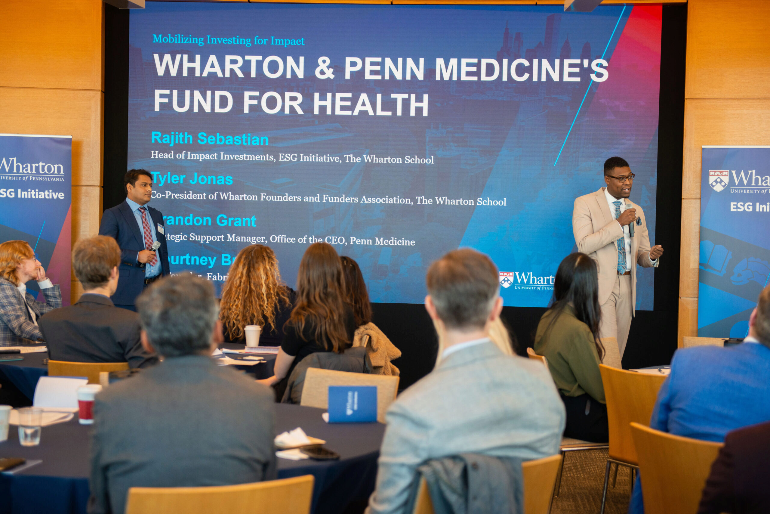 Brandon Grant and Rajith Sebastian in suits presenting to a room full of people in business attire in front of a slide that says "Wharton & Penn Medicine's Fund for Health"