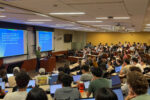 Image of a packed tiered classroom of students, some with laptops open, watching a speaker present research from the front of the classroom.