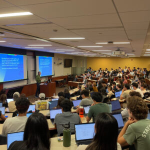 Image of a packed tiered classroom of students, some with laptops open, watching a speaker present research from the front of the classroom.