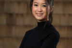 Marielle Kang posing for a professional headshot in a black turtleneck against a brick wall