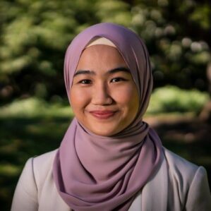 Sofea Shaifuddin smiling in a cream blazer and mauve hijab outside with green foliage in the background.