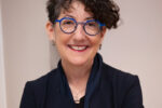 Suzanne Biegel posed smiling in a professional headshot wearing all black and bright blue glasses