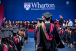 Wharton MBAs walking towards the stage at graduation in graduation regalia with students seated on both sides of the walkway
