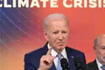 President Joe Biden stands in front of a microphone with his pointer finger up in front of a backdrop in sunset colors that says CLIMATE CRISIS in white letters and a man stands behind him.