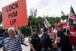 A group of police officers and protesters stand in a road; some protestors hold American flags and one holds a red sign that reads "LOCK HIM UP."
