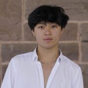 Photo of Raymond Liu Ao smiling towards the camera in a white button down shirt in from of a stone wall