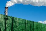 A smokestack with smoke billowing into the sky with a green wall in front of it.