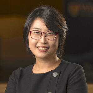 Danqing Zhu smiles in a black blouse and red glasses in front of a dark background.