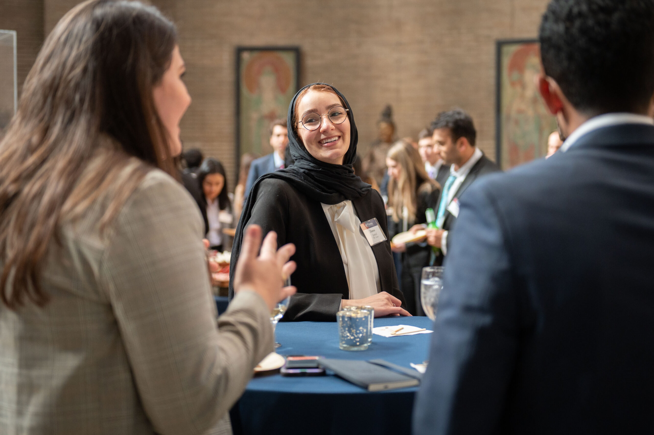 A group of students is smiling and talking at the evening reception. The shot focuses on one student smiling at a high table.