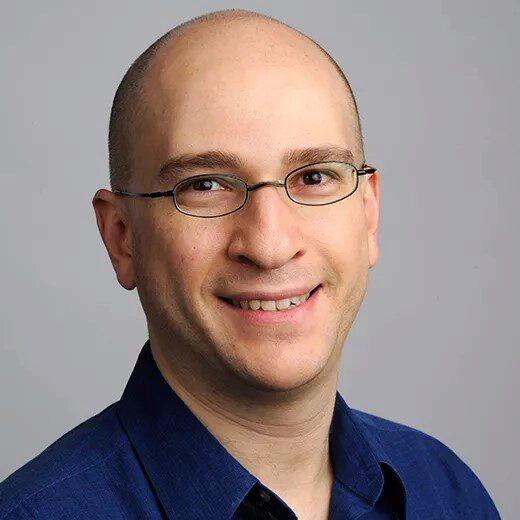 Yavok Bart smiles for a professional headshot in a navy blue button down in front of a grey background.