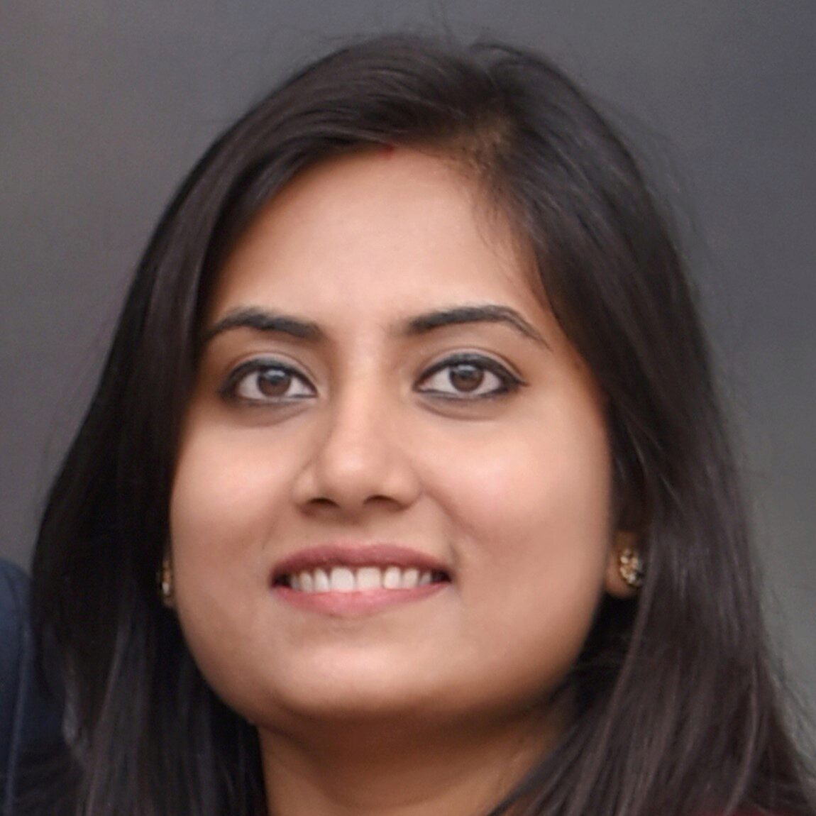 Sonam Singh smiles towards the camera in a close up photo in front of a dark grey background.