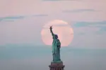 The statue of liberty in front of a super moon with blue clouds and a pink sky.