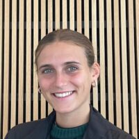 Headshot of a smiling person with short hair and hoop earrings, standing in front of a wooden slatted background.