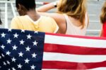 An American Flag with four people standing behind it, their backs to the camera.