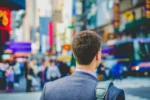 The back of a person with short brown hair wearing a blue blazer in front of a busy city backdrop.