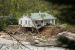 A house devastated by a flood with a tree fallen atop it and the ground eroded underneath it.