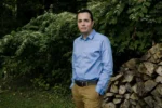 Zeke Hernandez stands in slacks and a blue button down shirt in front of a stack of wood and a tree.