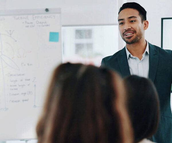 A man in a suit presents to a classroom with a large pad with writing in the the background.