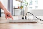 A hand pushes the button on a power strip that has five cords plugged into it in the backdrop of an apartment.