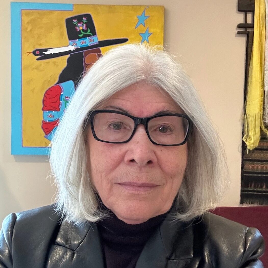 Rebecca Adamson smiles towards the camera in a black leather coat in front of artwork.