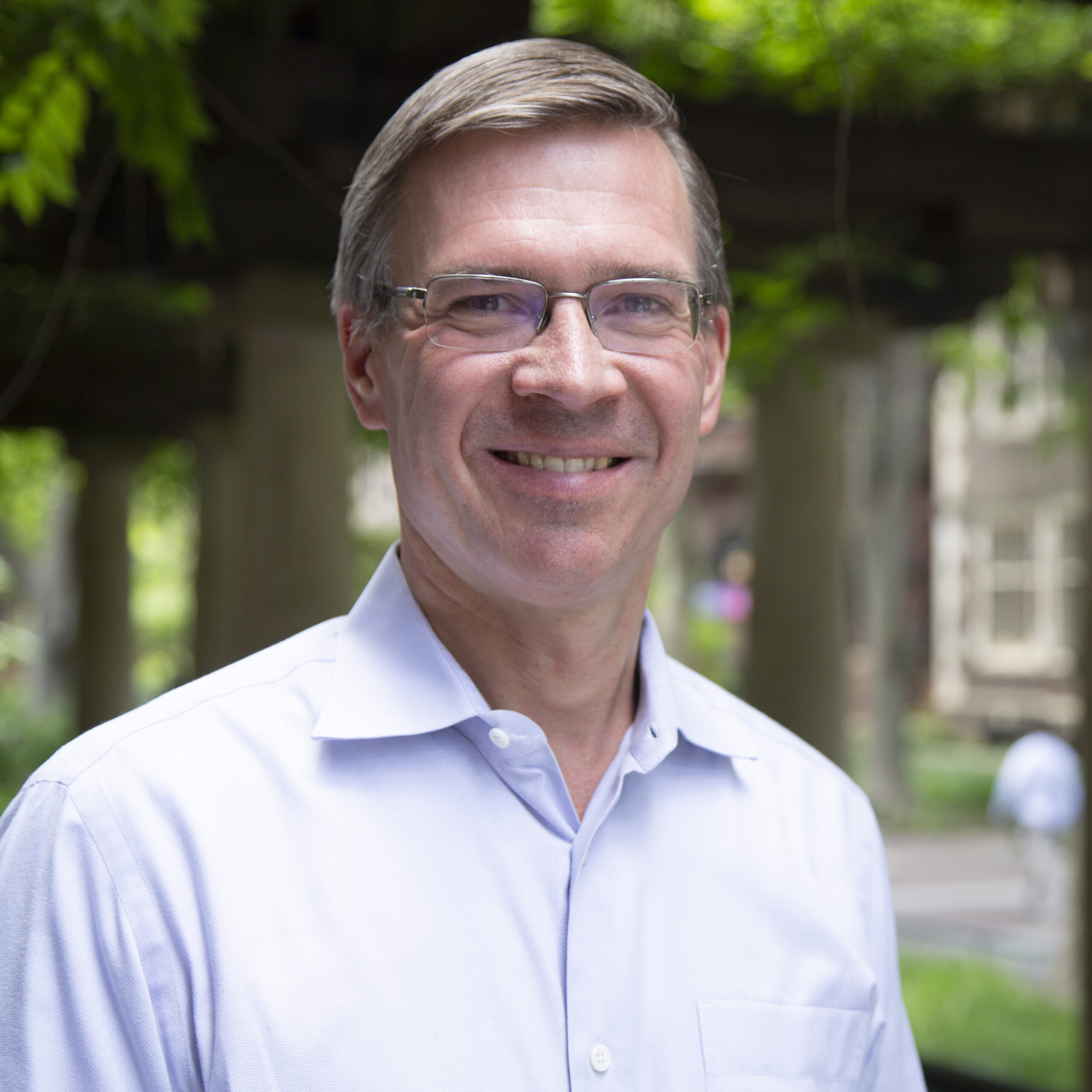 Witold Henisz smiles towards the camera in a light blue button-down shirt in front of trees.