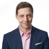 Eli Emigh smiles into camera wearing a pink button down shirt and a navy blazer.